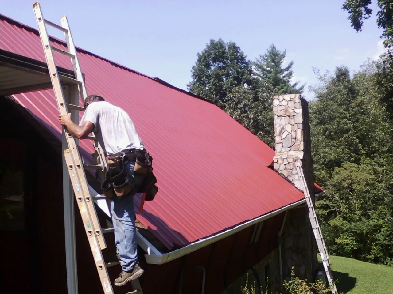 Roofer on a ladder beside a red metal roof and stone chimney — Soffit & Fascia Repair by Roofing Brothers in Shubuta